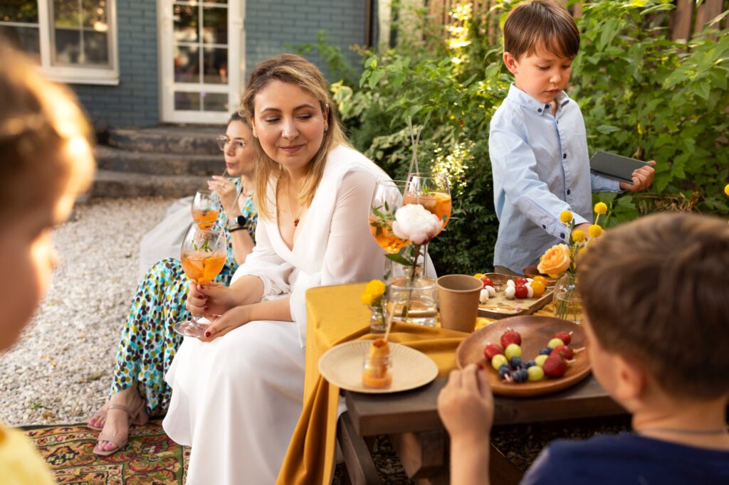 Happy brunette female guest and bride drinking coctails by wedding table, spending time with kids